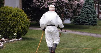 Pesticide technician walking on mowed lawn and dragging a long yellow hose behind him. He's fully covered by a white uniform, high black boots, black gloves and white hat.