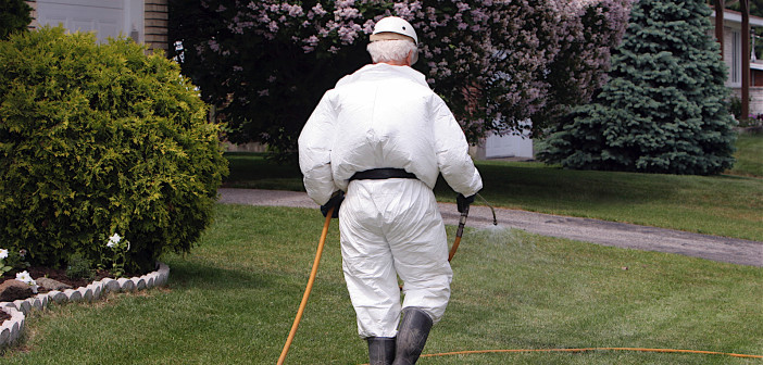 Pesticide technician walking on mowed lawn and dragging a long yellow hose behind him. He's fully covered by a white uniform, high black boots, black gloves and white hat.