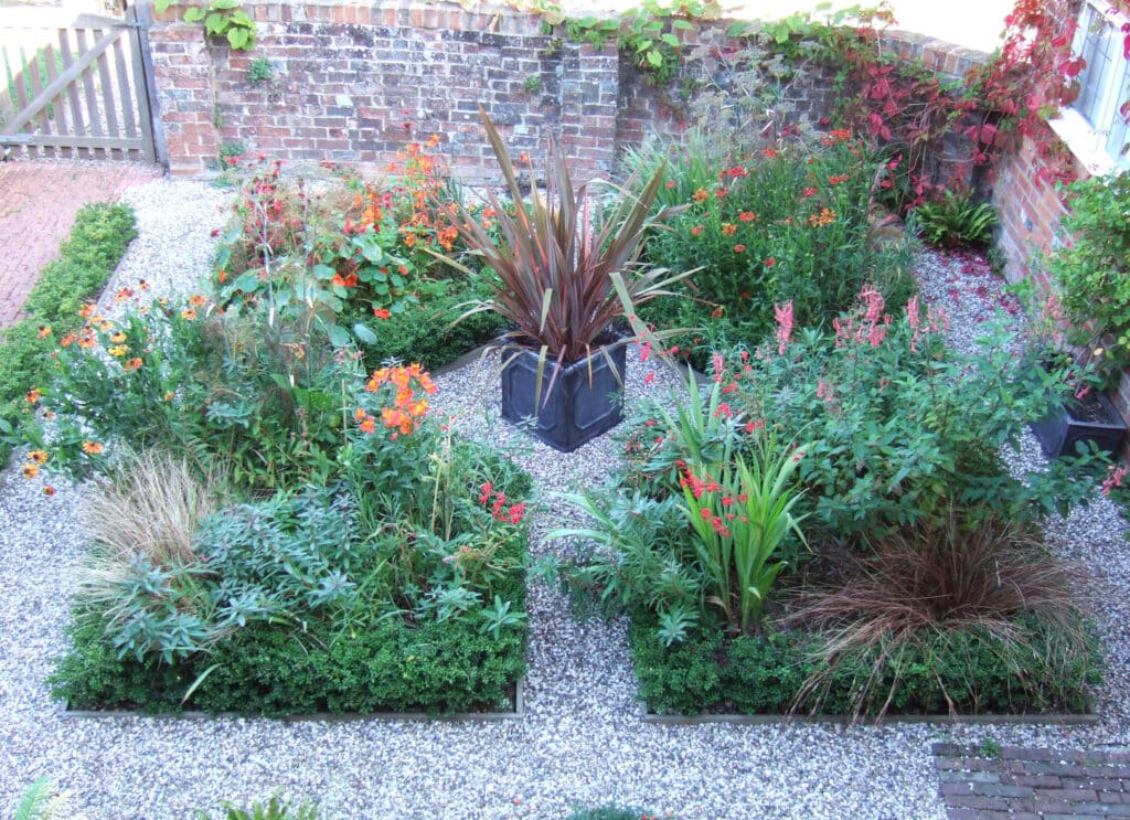 A small courtyard has four square groups of flowers surrounded by gravel.