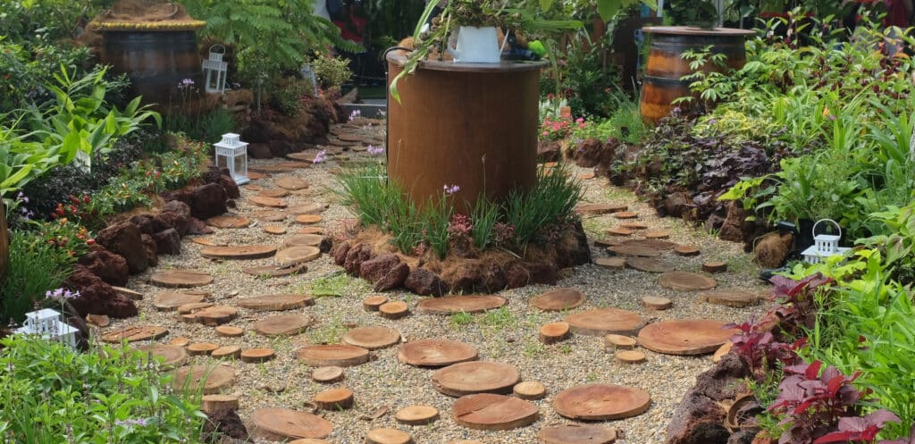 A garden path surrounded on each side by plants. It splits in the middle to go around a large brown barrel.